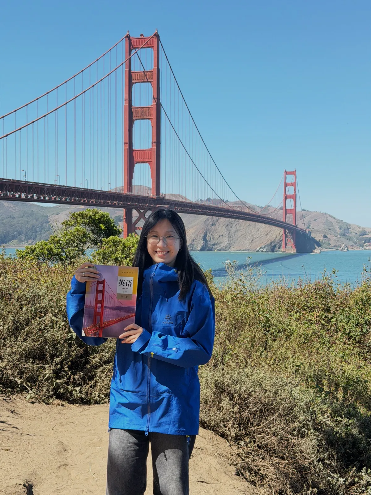 Yijing standing in front of the Golden Gate Bridge, holding up a Chinese textbook whose cover shows the same bridge.