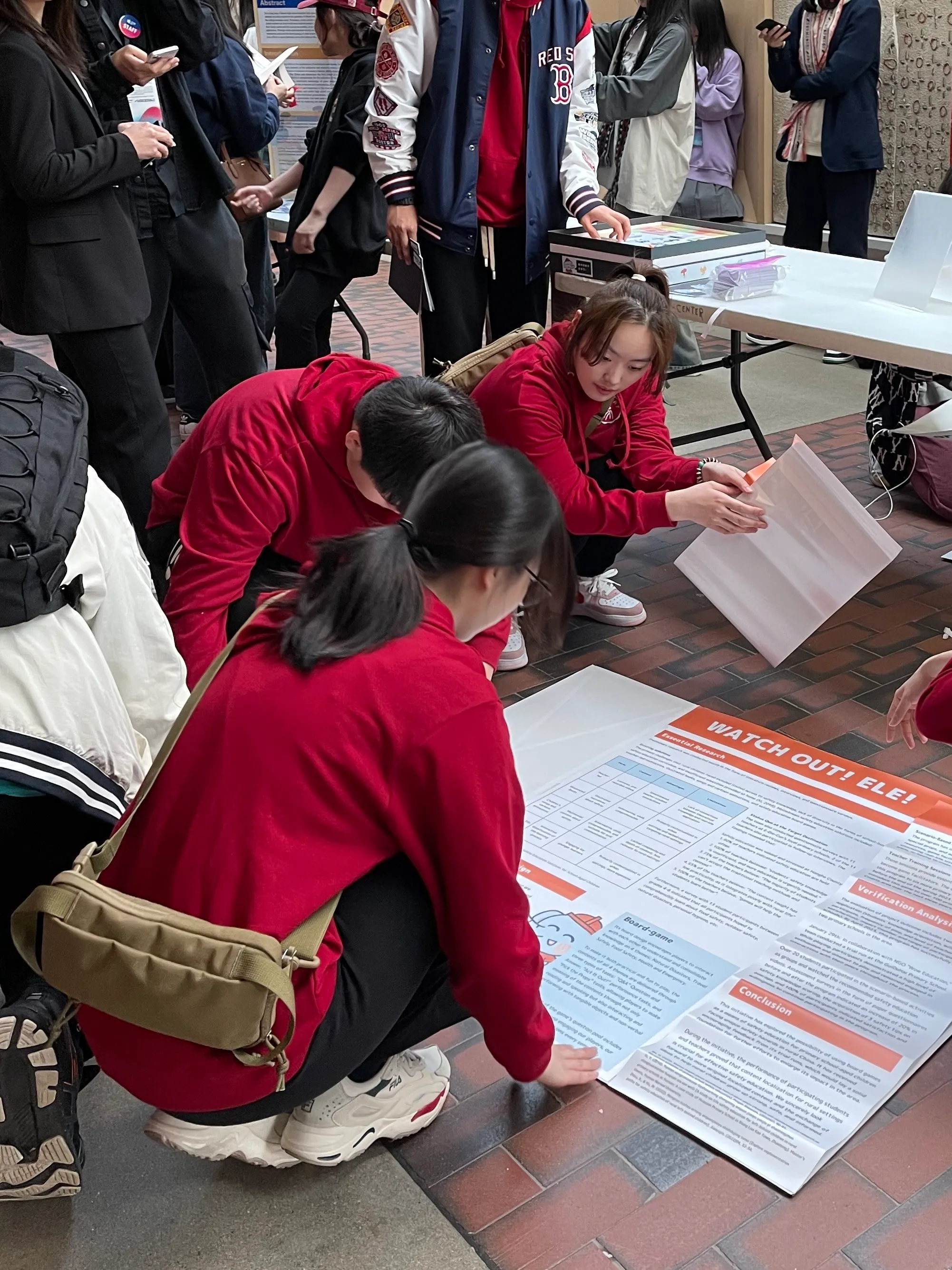 Team members in red hoodies crouching on a brick floor to lay out a large orange Watch Out! Ele! poster at a crowded exhibition.