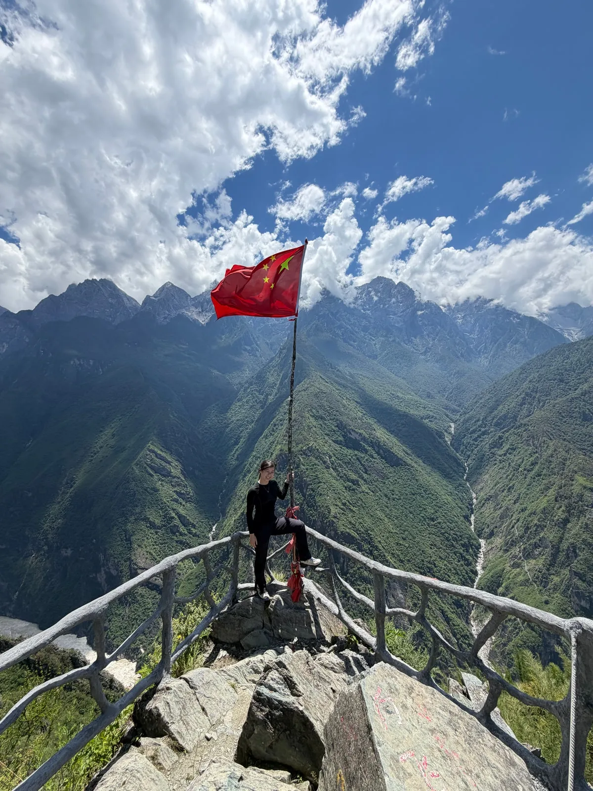Yijing sitting on the railing of a rocky mountain lookout beside a red flag, above a deep green gorge under a bright, cloudy sky.
