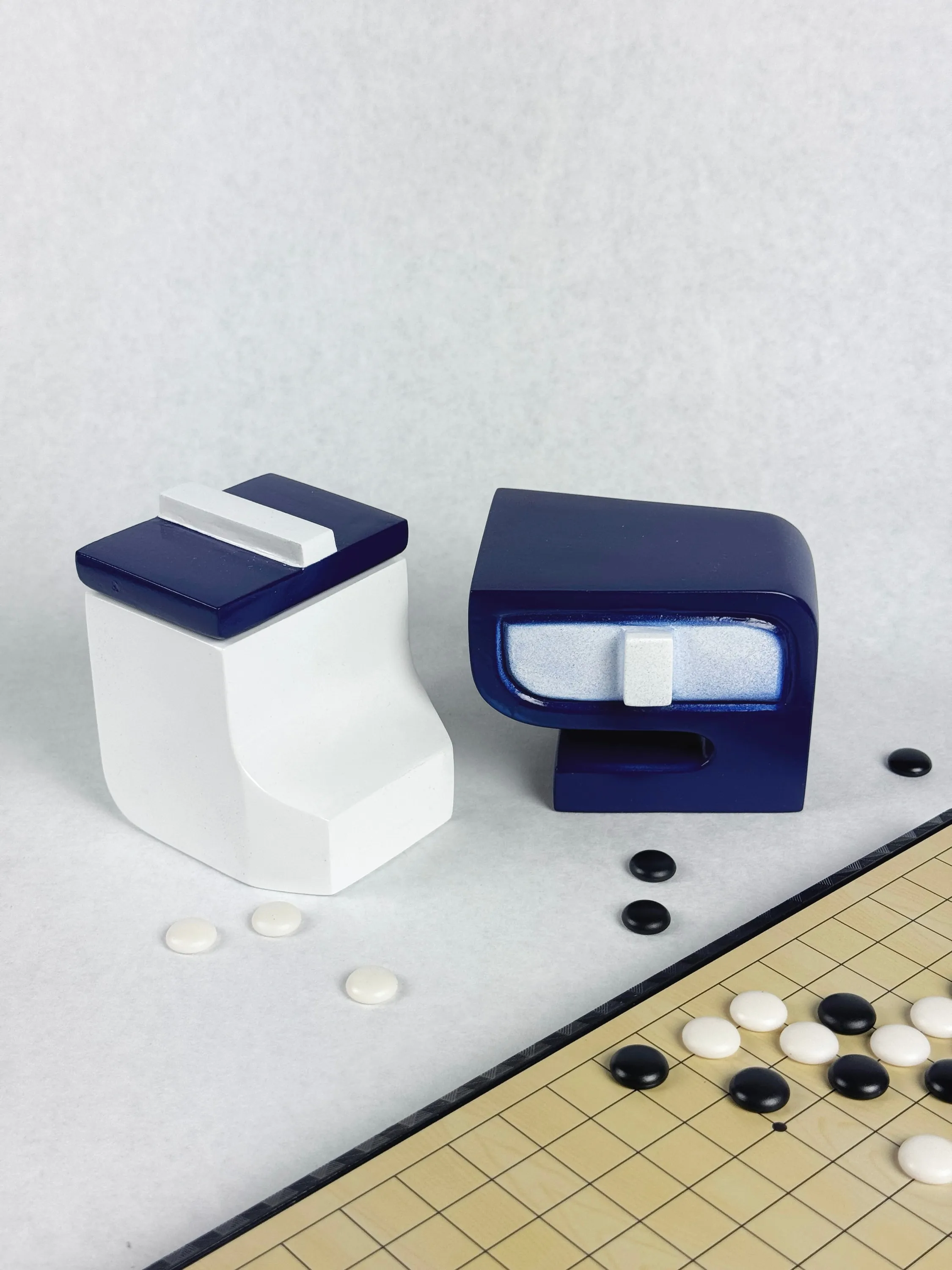 Two white and navy containers, one holding white Go stones, beside a Go board scattered with black and white stones.